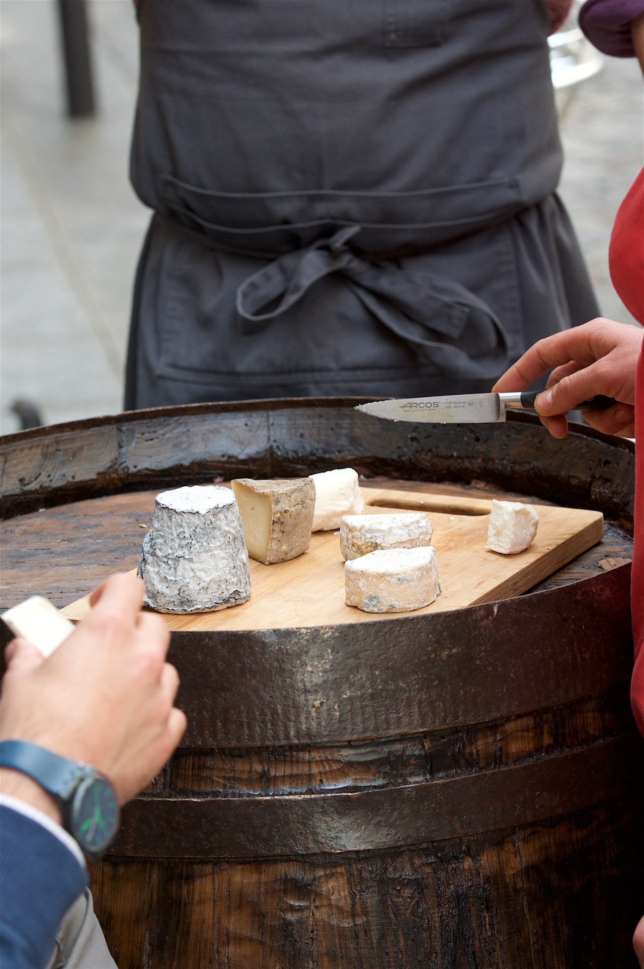 Person holding an Arcos knife cutting cheese samples arranged on wooden board in rustic barrel.