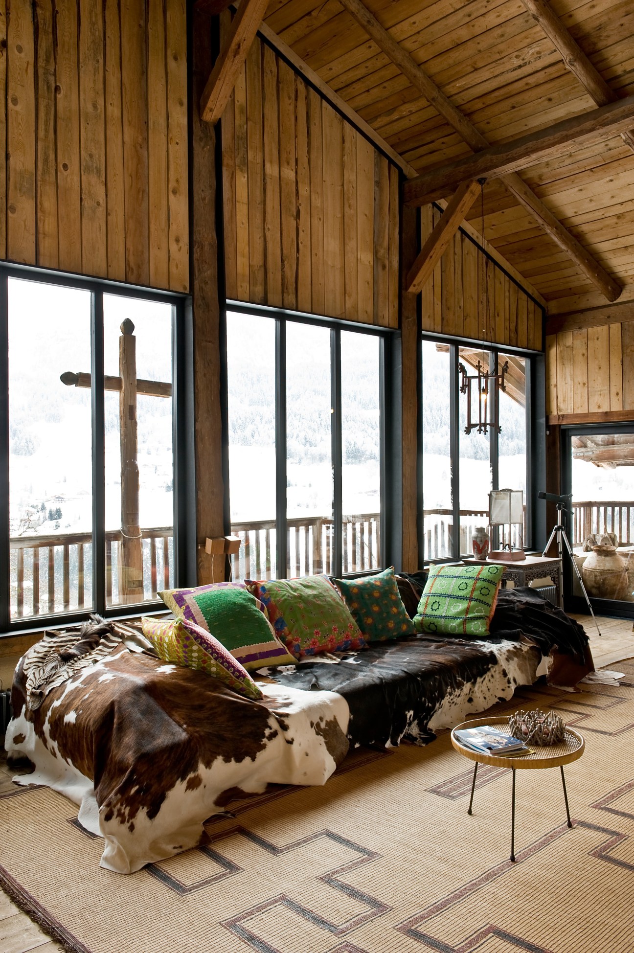 A cowhide-upholstered sofa with colorful patterned pillows beside floor-to-ceiling windows overlooking snowy mountains.