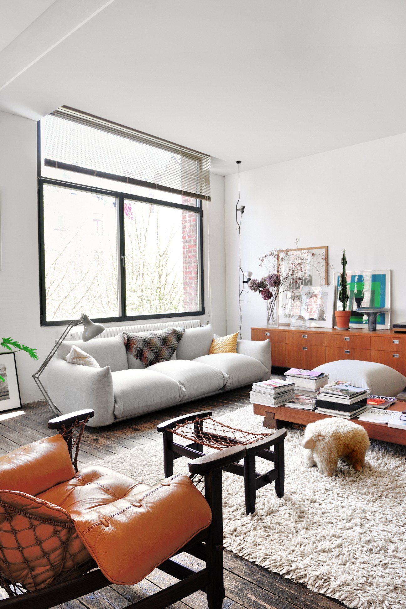 Modern living room with grey Arflex sofa, orange leather chair, wooden furniture, and a dog on a white rug.