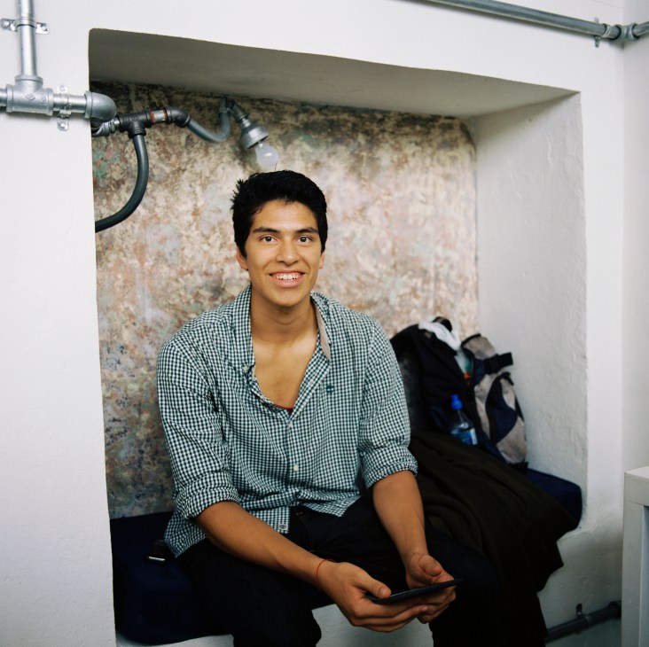 Young man in checkered shirt smiling at camera in a sparse room with exposed brick wall and plumbing fixtures.