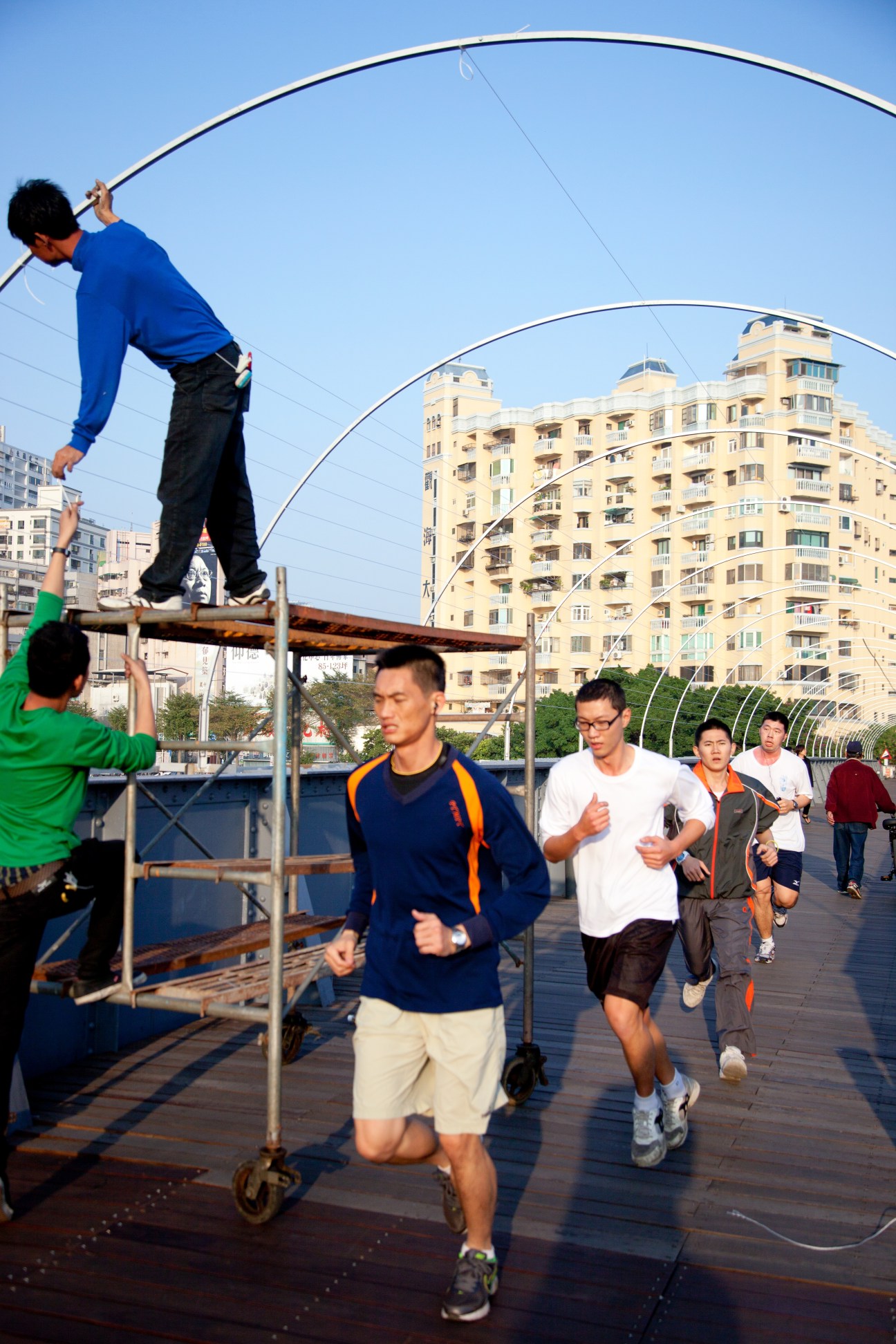 People jogging and exercising on a waterfront path with a child jumping rope overhead, apartment buildings in background.