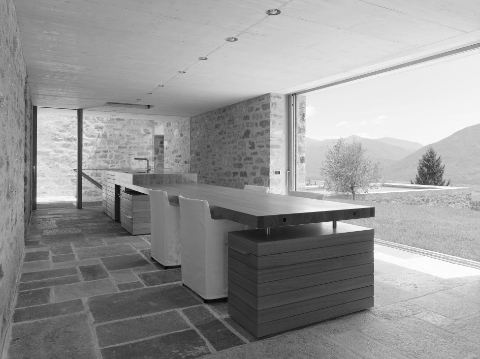 Modern minimalist interior with stone walls, dining table, and panoramic mountain views through large windows.