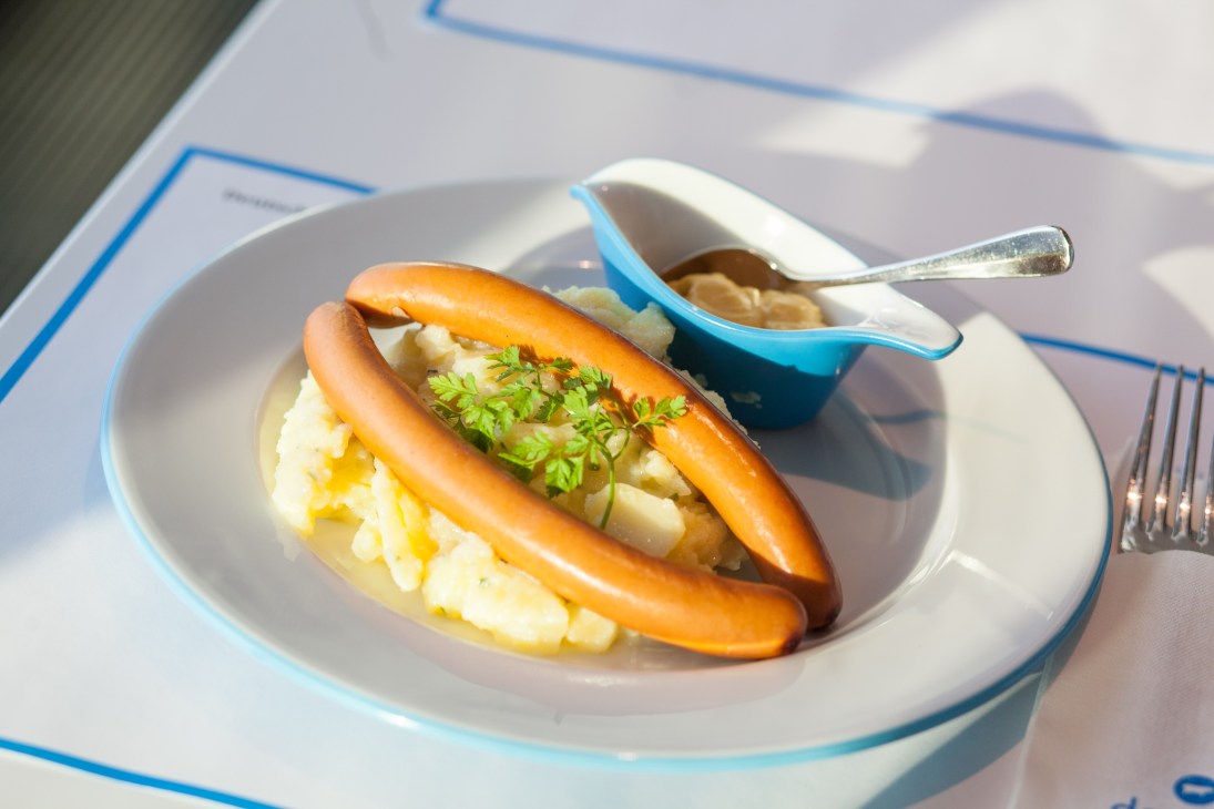 A sausage sandwich with mustard and parsley garnish on a white plate with blue trim at Deutsch restaurant.