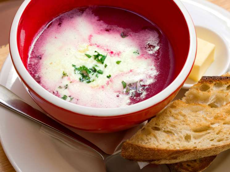 A red and white bowl of beet soup with cream and chives, served with toasted bread.