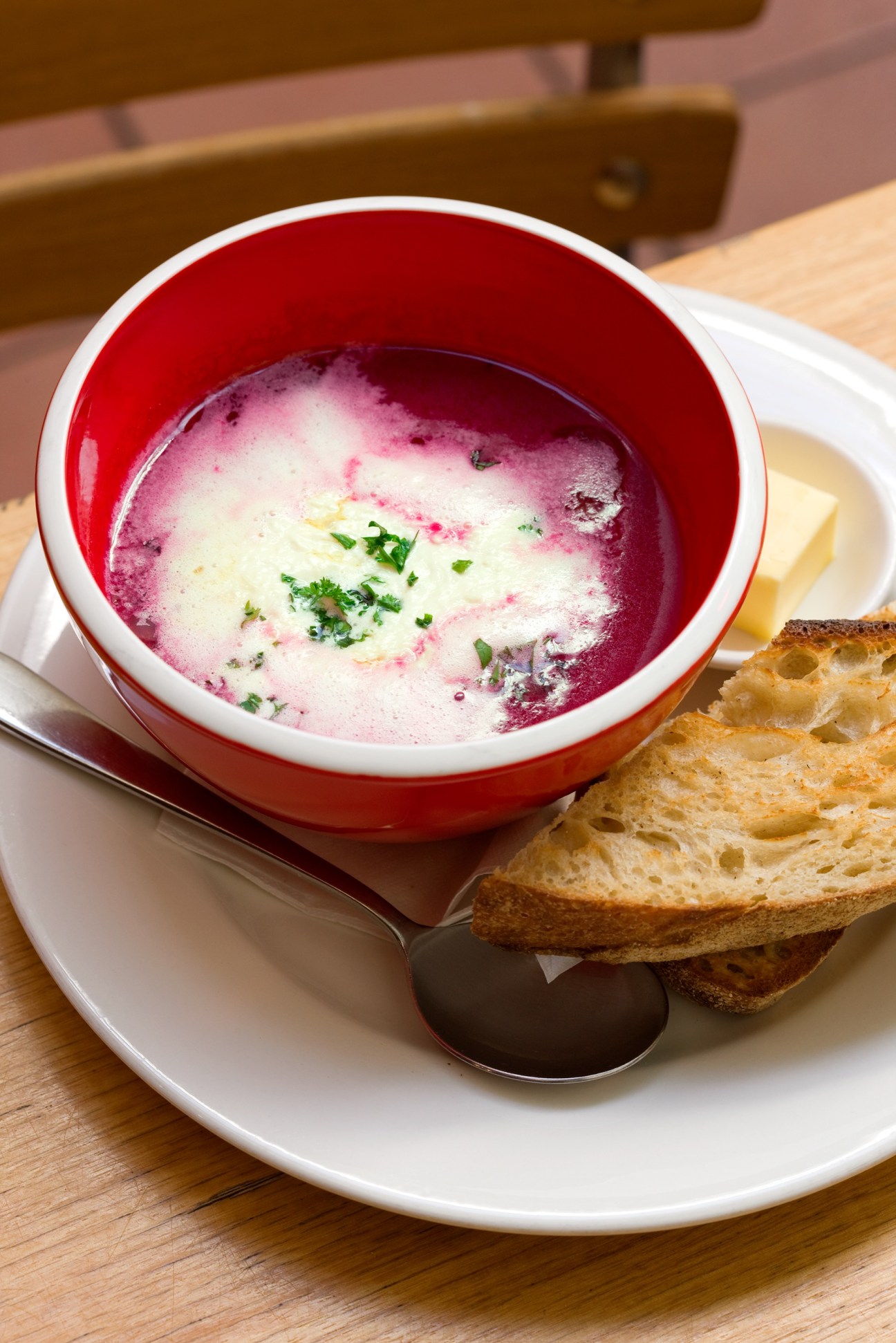 A red and white bowl of beet soup with cream and chives, served with toasted bread.