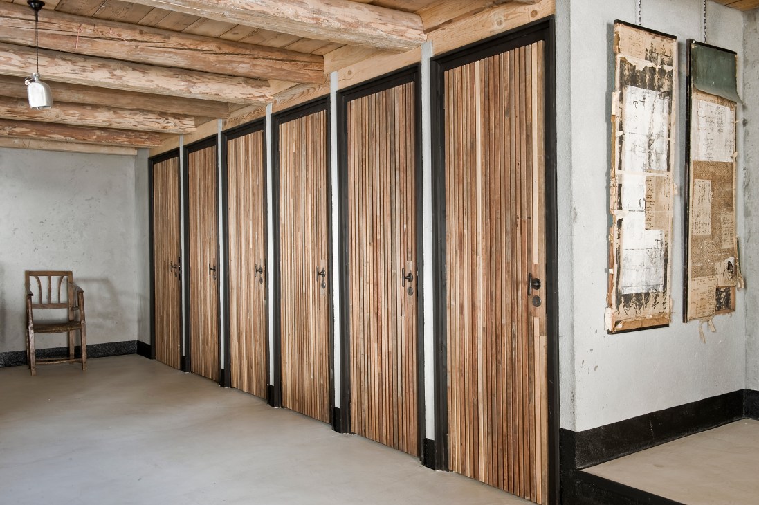Series of wooden storage cupboards with vertical slat doors and black frames line a white wall beneath exposed timber beams.
