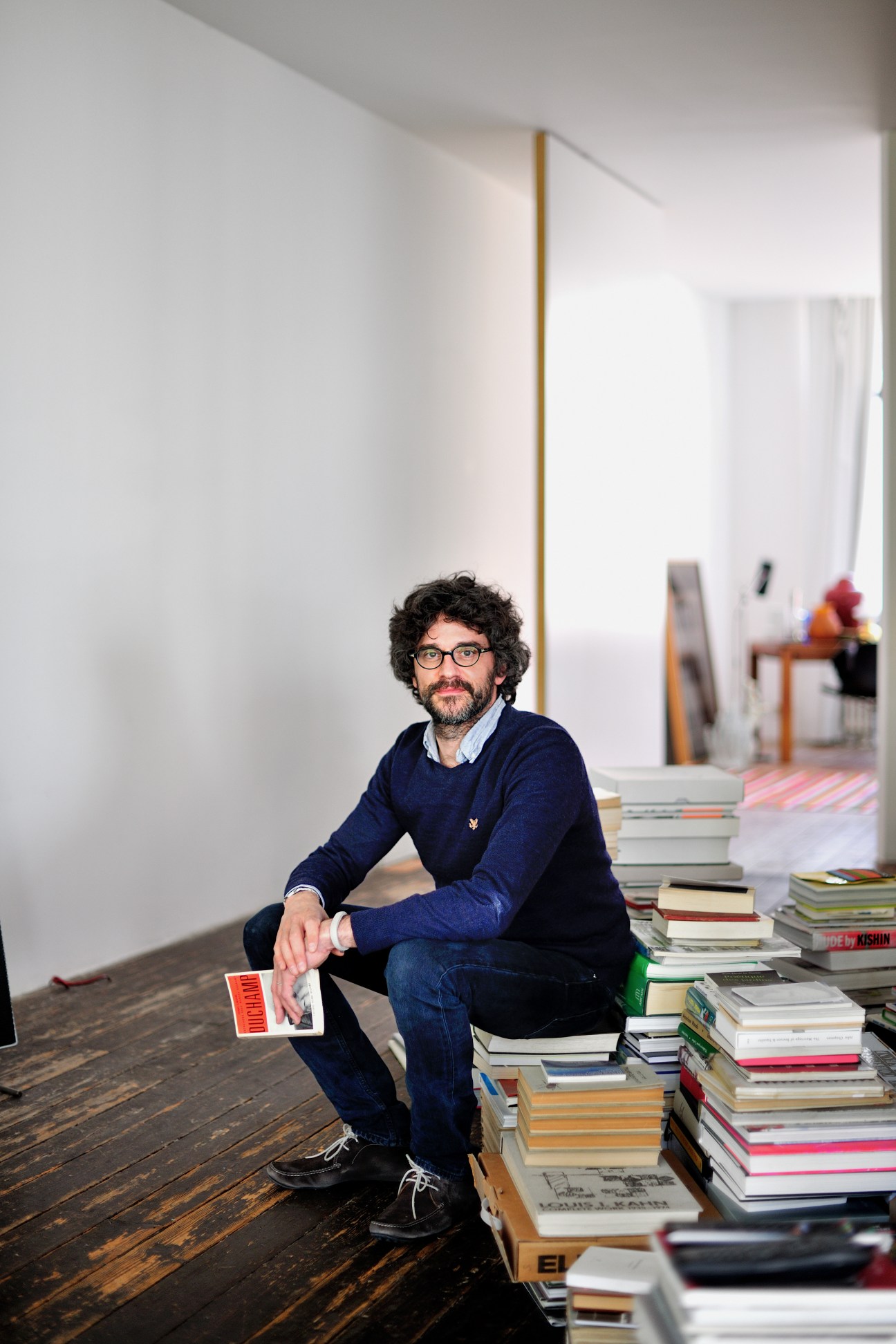 Pierre Lhoas, architect, crouching among stacks of books in a bright studio space.