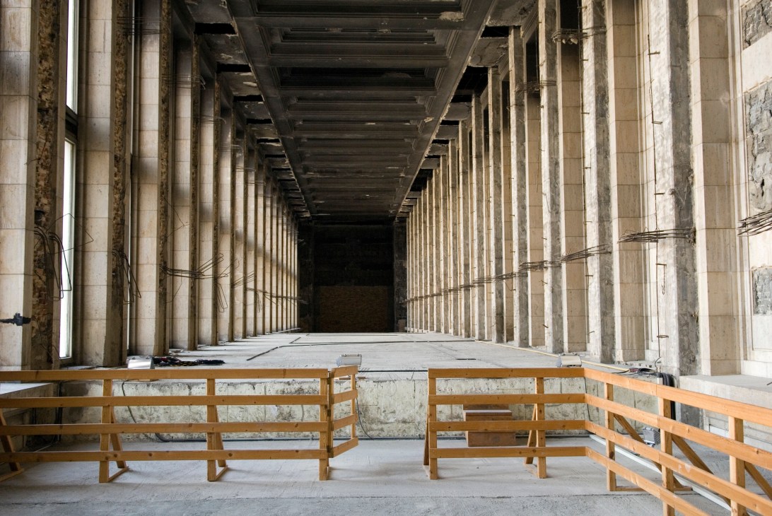 Interior of Tempelhof airport's Ruhmeshalle with wooden benches facing a long colonnade of stone pillars.