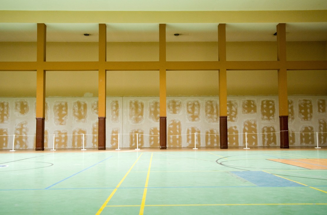 Empty sports hall with wooden columns, yellow markings on floor, and large windows showing shadows of buildings outside.