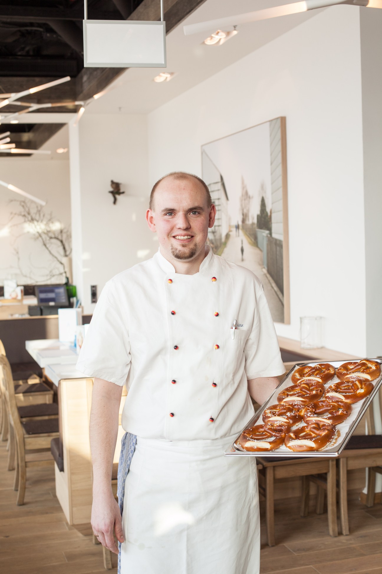 Chef in white double-breasted jacket holding tray of pretzels in modern restaurant.