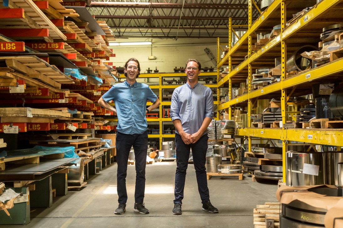 Two men stand smiling in a warehouse surrounded by yellow shelving units stocked with furniture parts and materials.