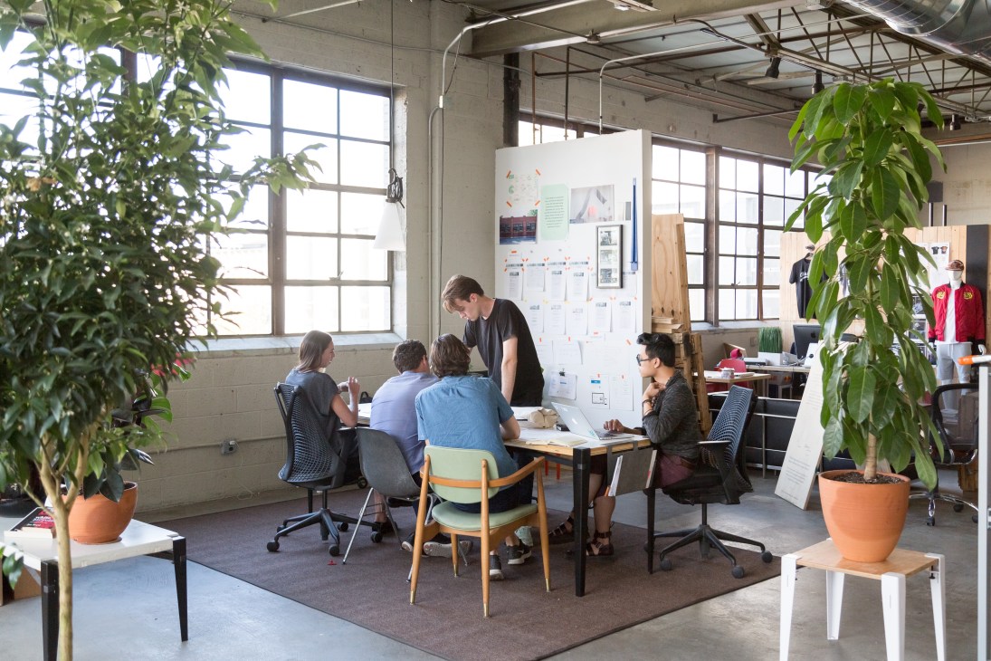 Floyd team members collaborate around a table in their Detroit office with industrial windows and potted plants.