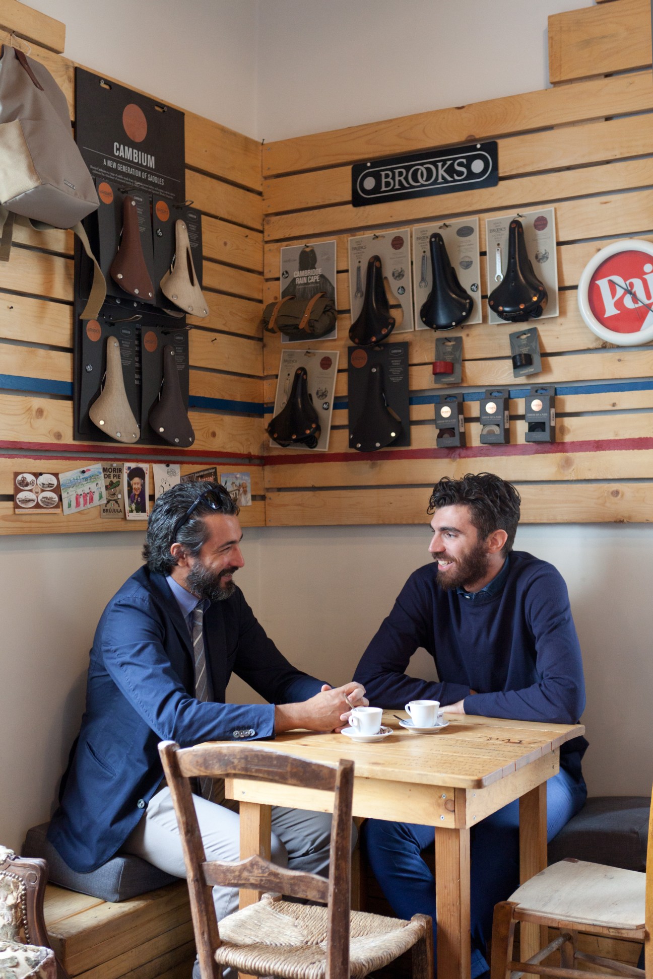 Two men sit at a wooden table with coffee cups at Pai Bikery, with bicycle saddles and accessories displayed on the wall behind them.