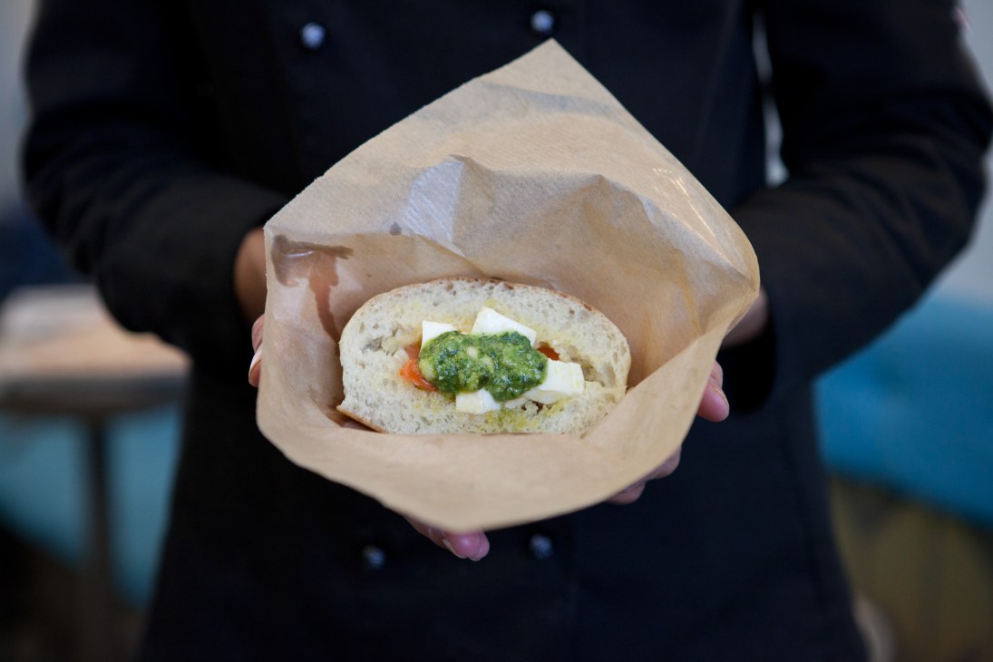 A person in a black coat holds a paper-wrapped panini sandwich filled with pesto, cheese and vegetables.