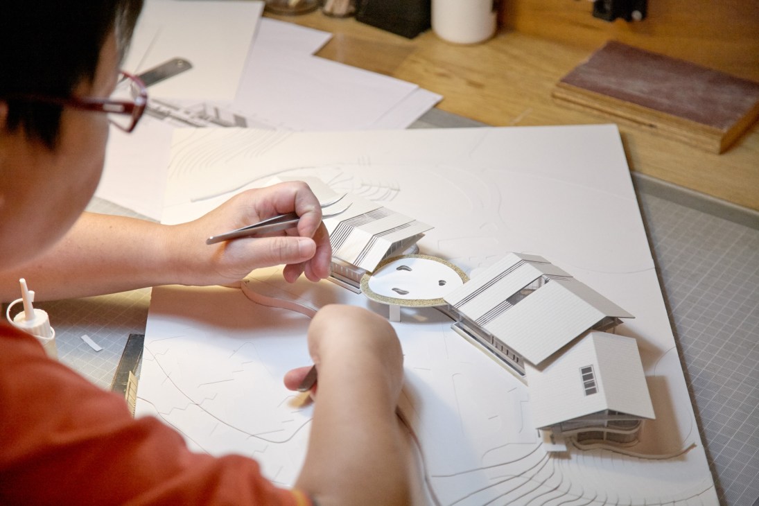 Person working on architectural drawings and miniature model with design tools on wooden desk.