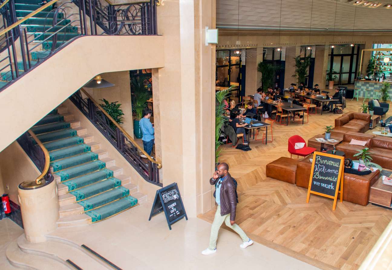 Overhead view of WeWork's art deco lobby with workers at wooden tables, leather sofas, and geometric glass ceiling in Paris.