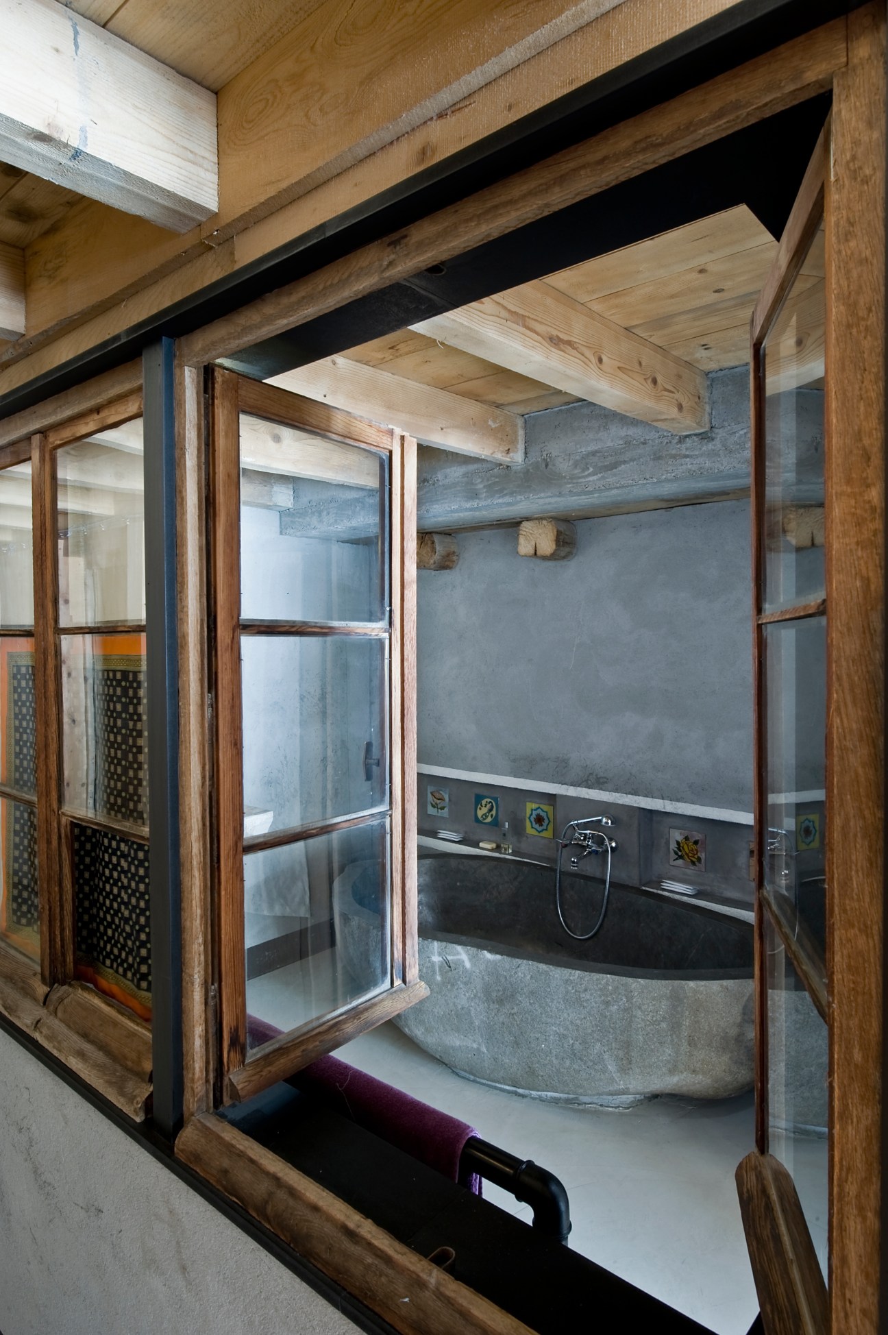 Stone bathtub carved from rock with black fixtures visible through wooden-framed glass enclosure.