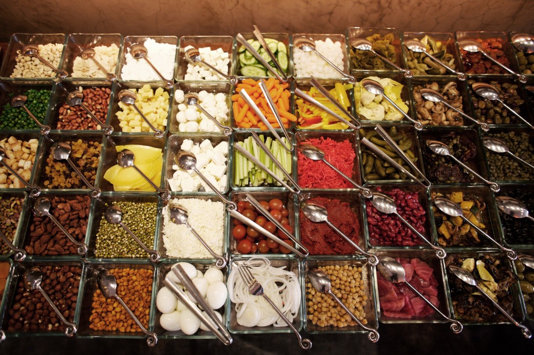 Saladicious restaurant's extensive salad bar with numerous glass containers of fresh vegetables, legumes, and toppings with serving utensils.