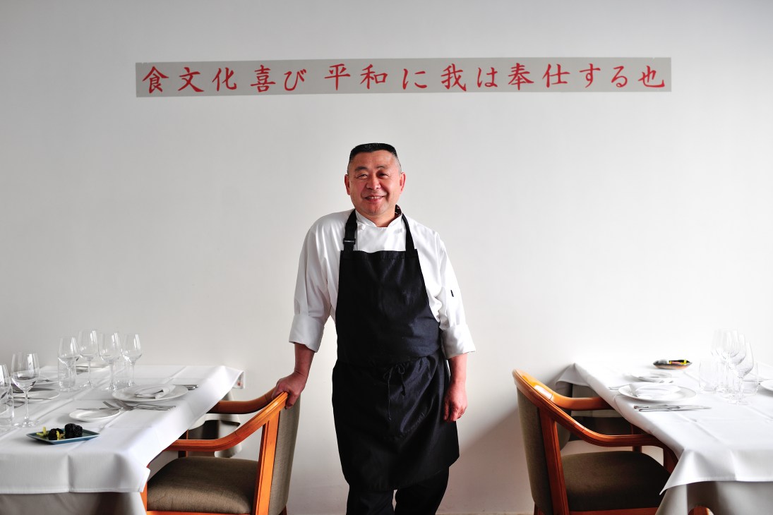 Chef Saburo Inada standing in his restaurant dining room wearing a black apron and white shirt.