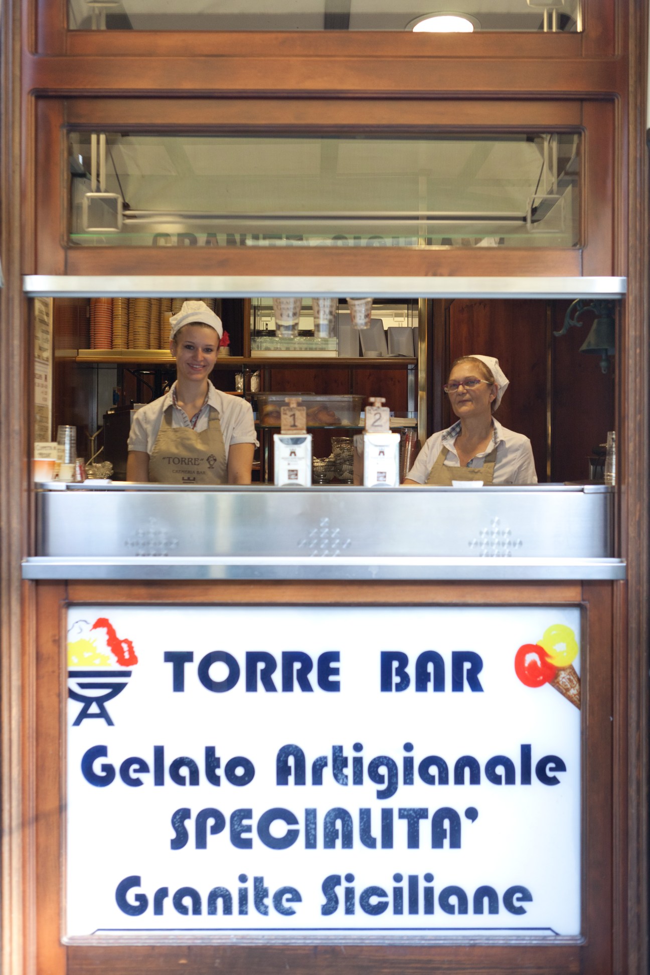 Two staff members inside Torre Bar gelateria shop window with signage advertising gelato artisanale and granite siciliane.