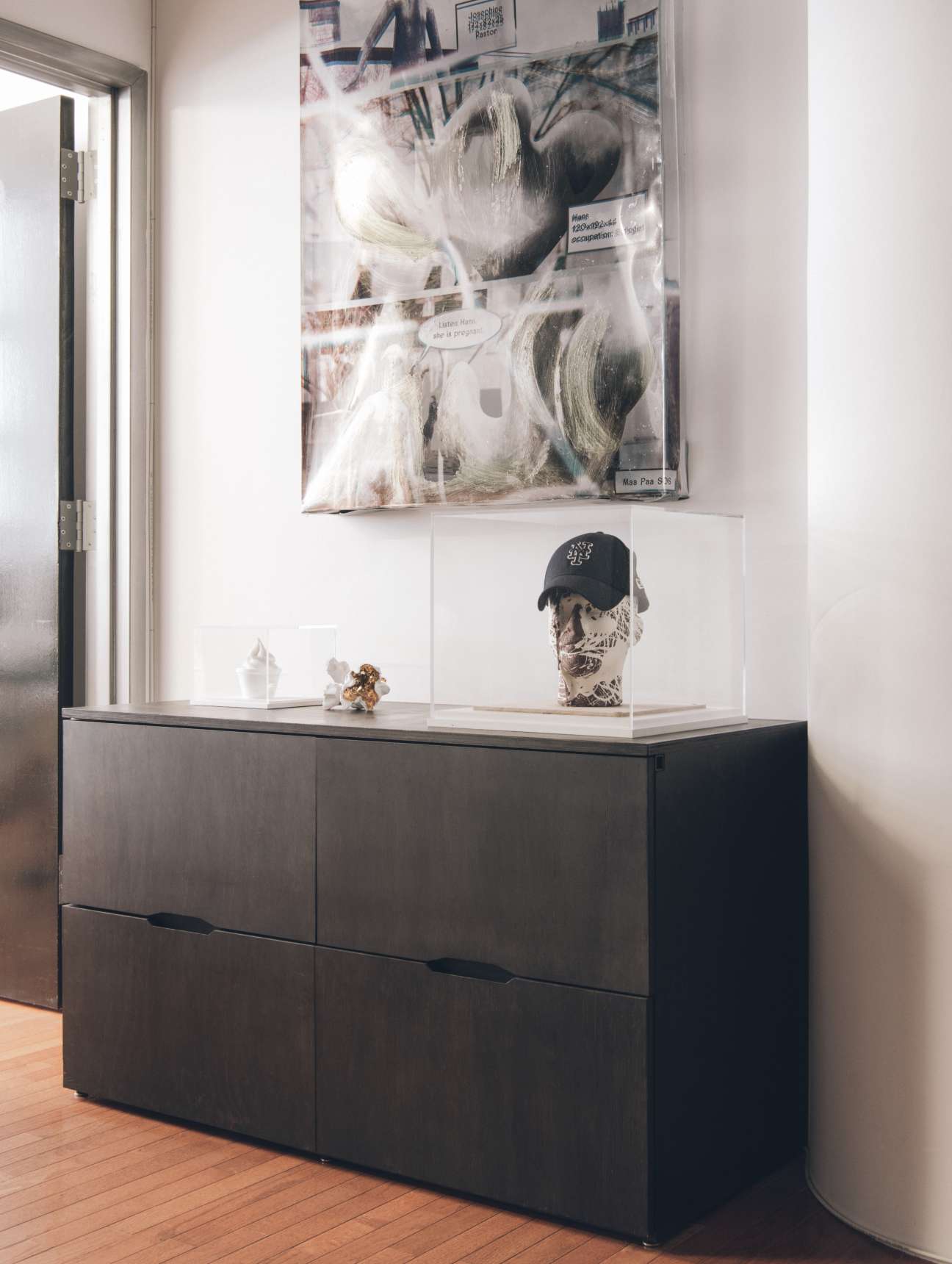 Contemporary artworks displayed on white shelving in a gallery space, including a framed print above and sculptural pieces below.