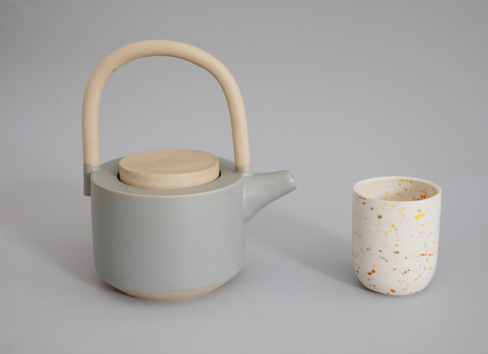 Gray ceramic teapot with wooden handle next to white cup with colorful speckled pattern on neutral background