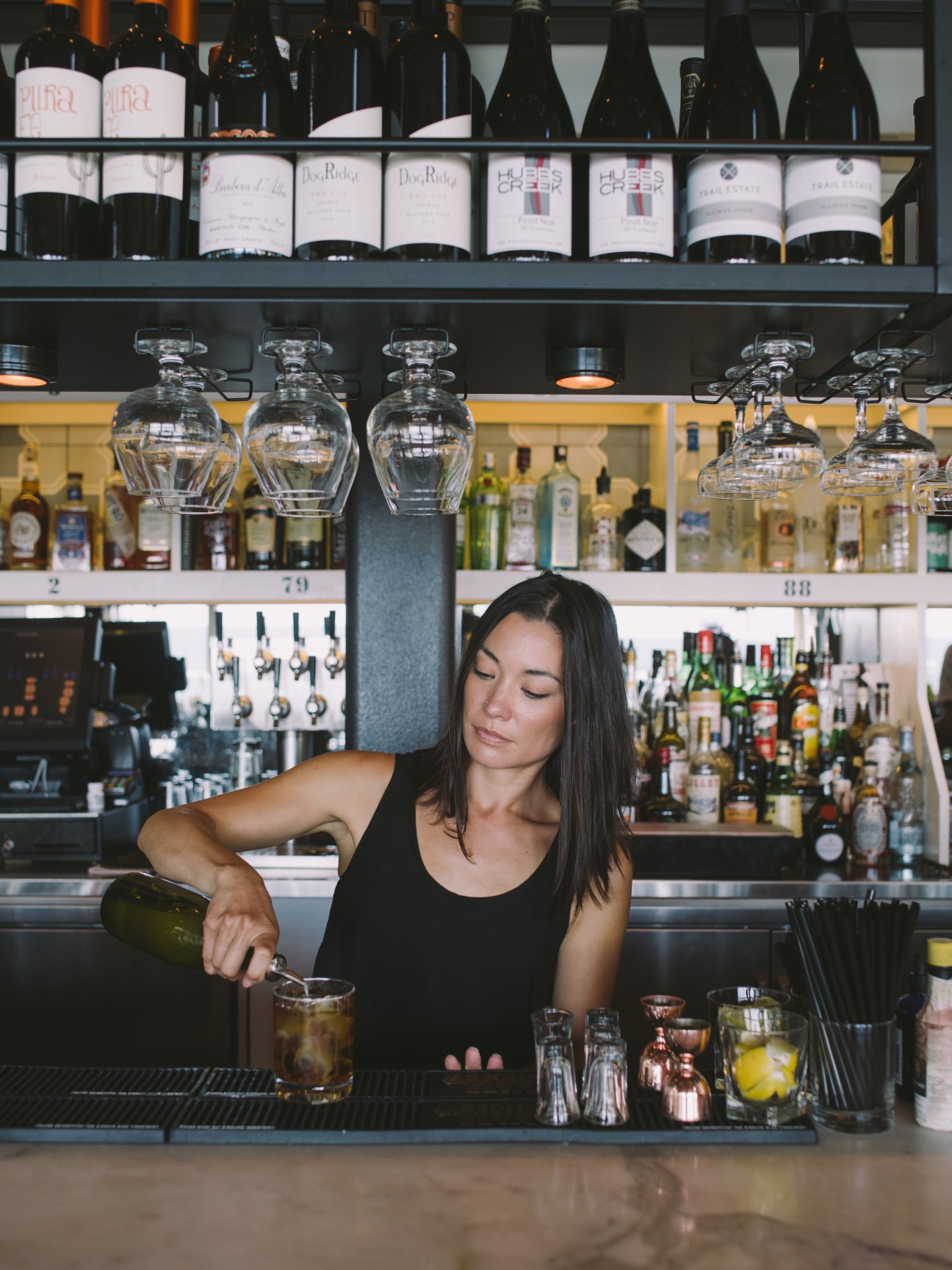 Female bartender in black top pouring cocktail behind bar with wine bottles and hanging glasses at Drake Devonshire Inn