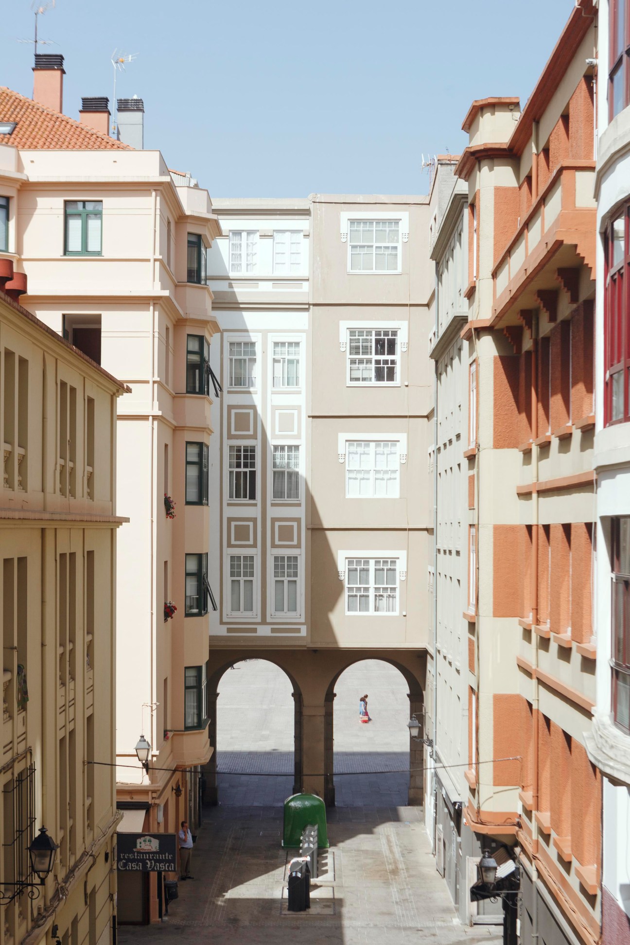 Narrow cobblestone street in A Coruña lined with colorful buildings framing an archway with ocean view beyond.