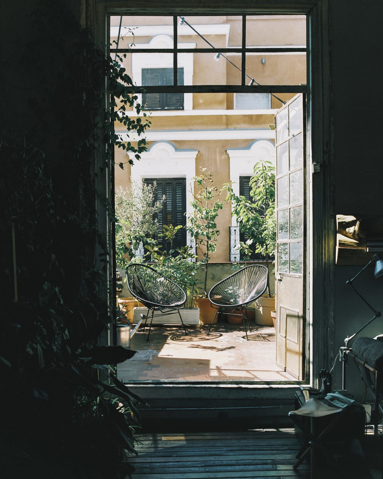 View through open doors to a sun-filled courtyard terrace with potted plants and two wire chairs