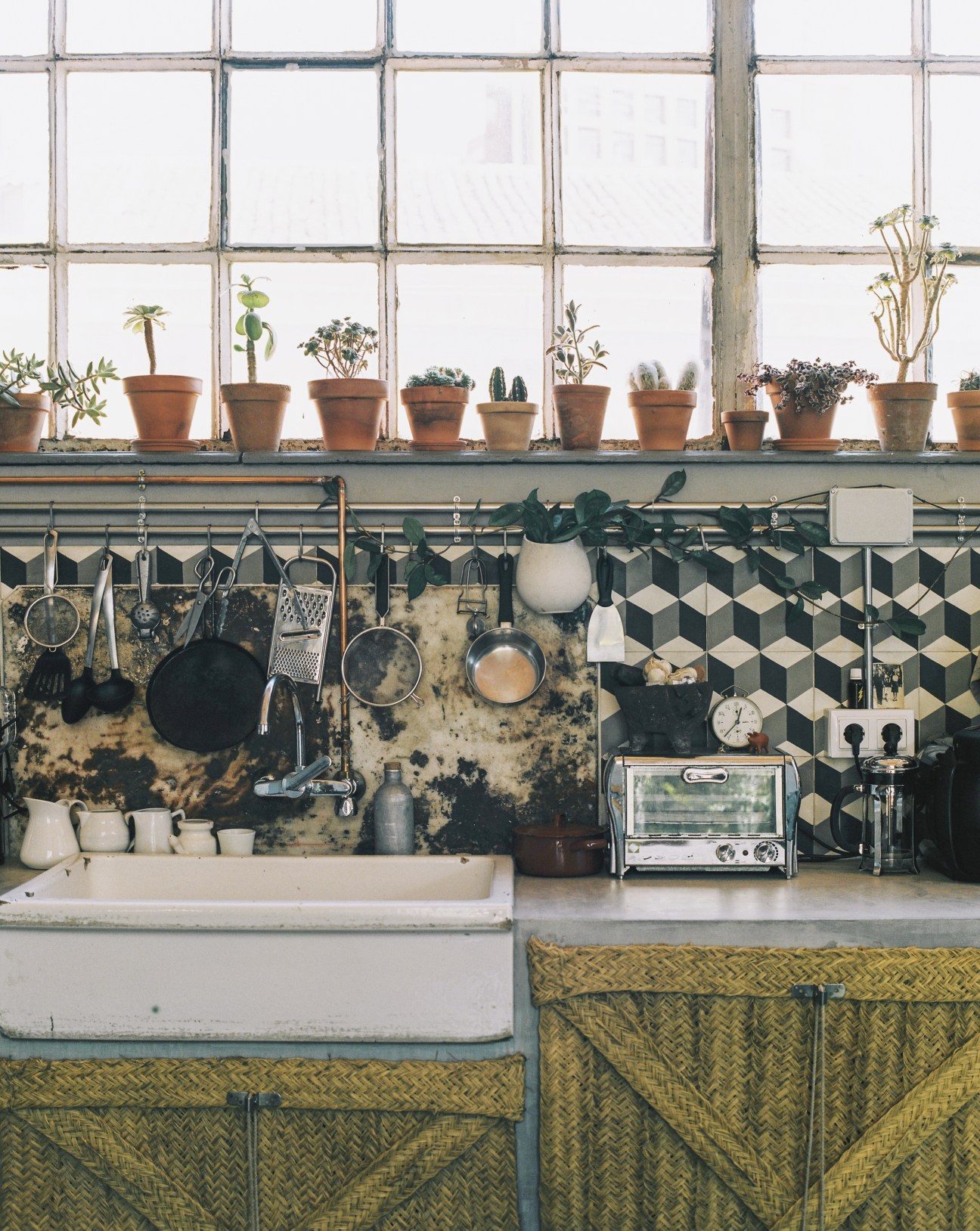 Kitchen workspace with potted plants on windowsill, hanging cookware, and geometric patterned backsplash.