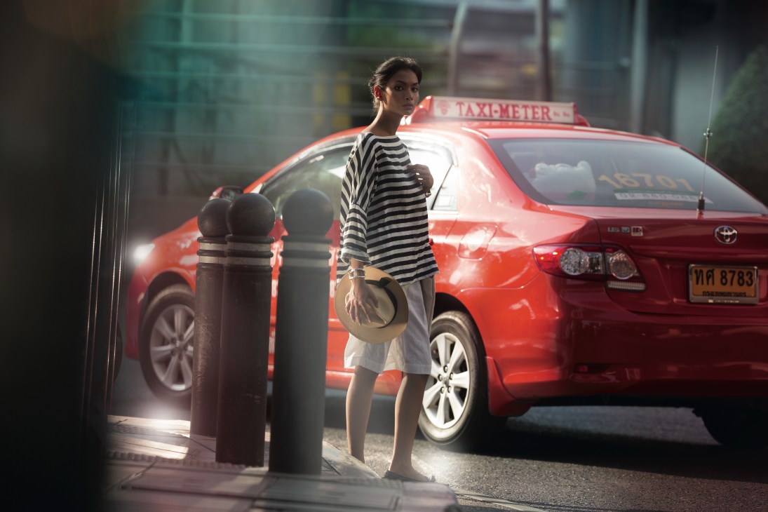 Woman in striped shirt and light trousers standing by bollards in front of a red Bangkok taxi.