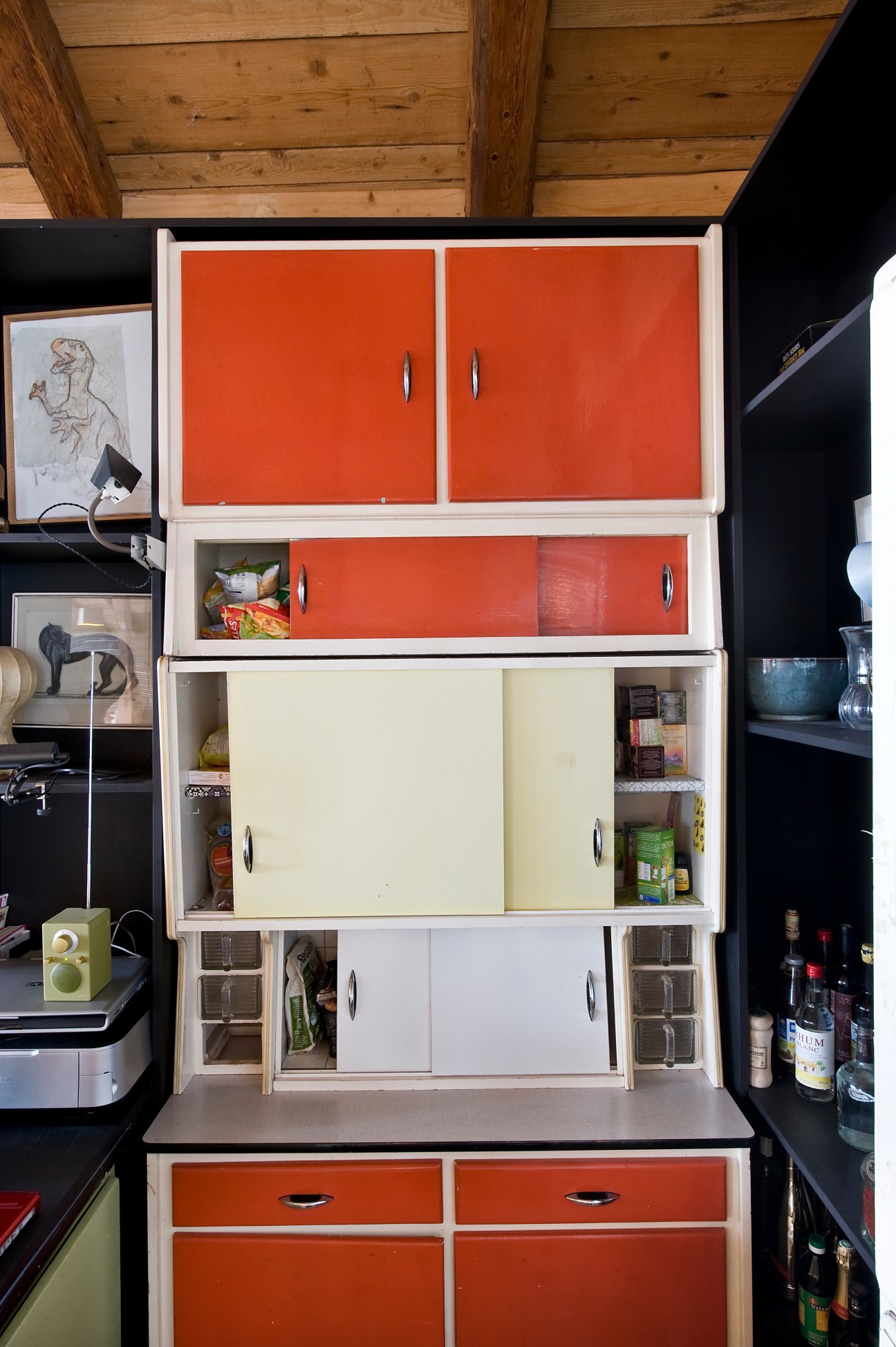 Tall modular kitchen storage unit with orange and cream-colored cabinet doors, open shelving displaying food items and kitchenware.