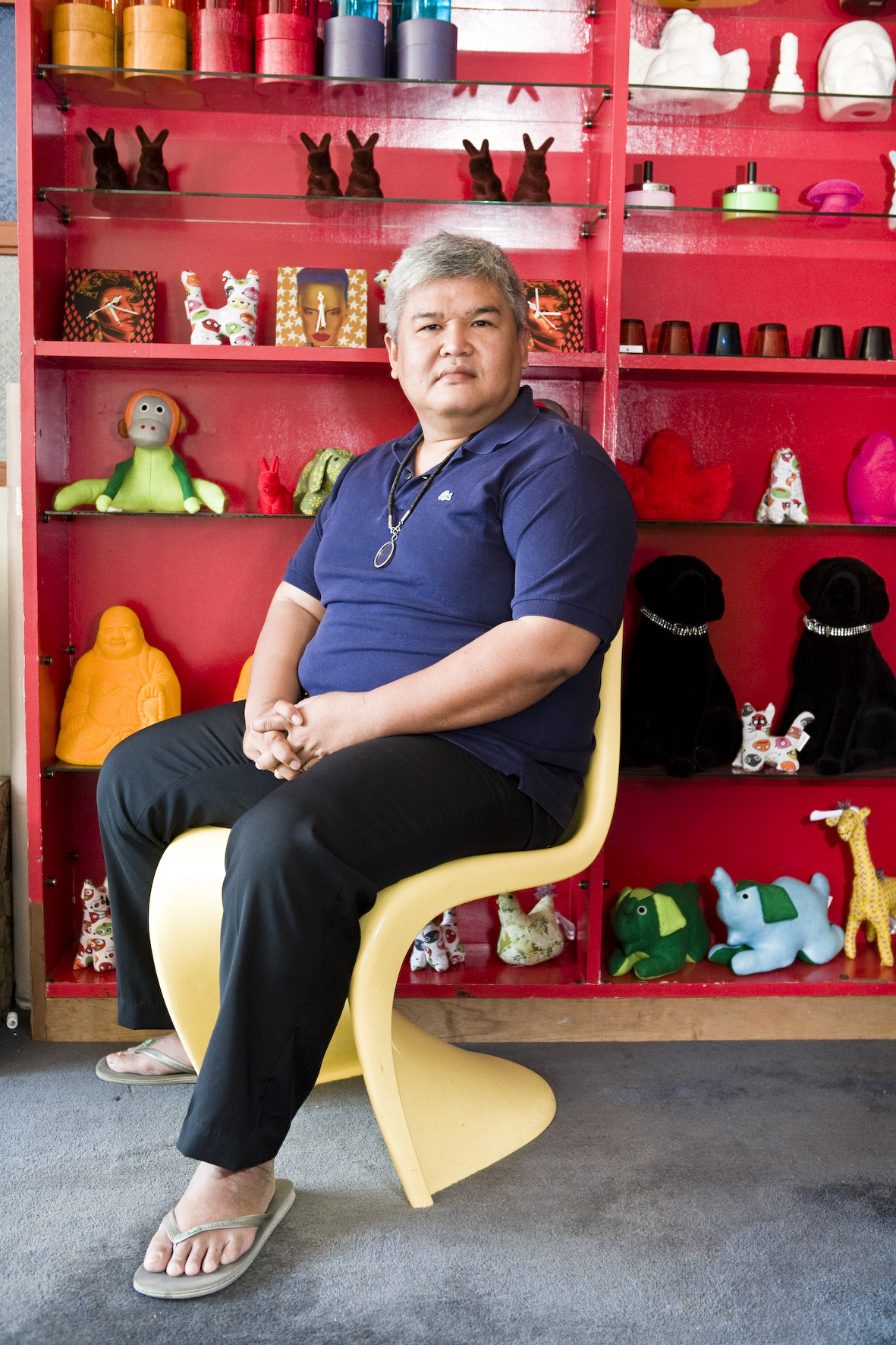 Anusorn Ngernyuang sits in front of a vibrant red shelving unit filled with decorative objects and collectibles.