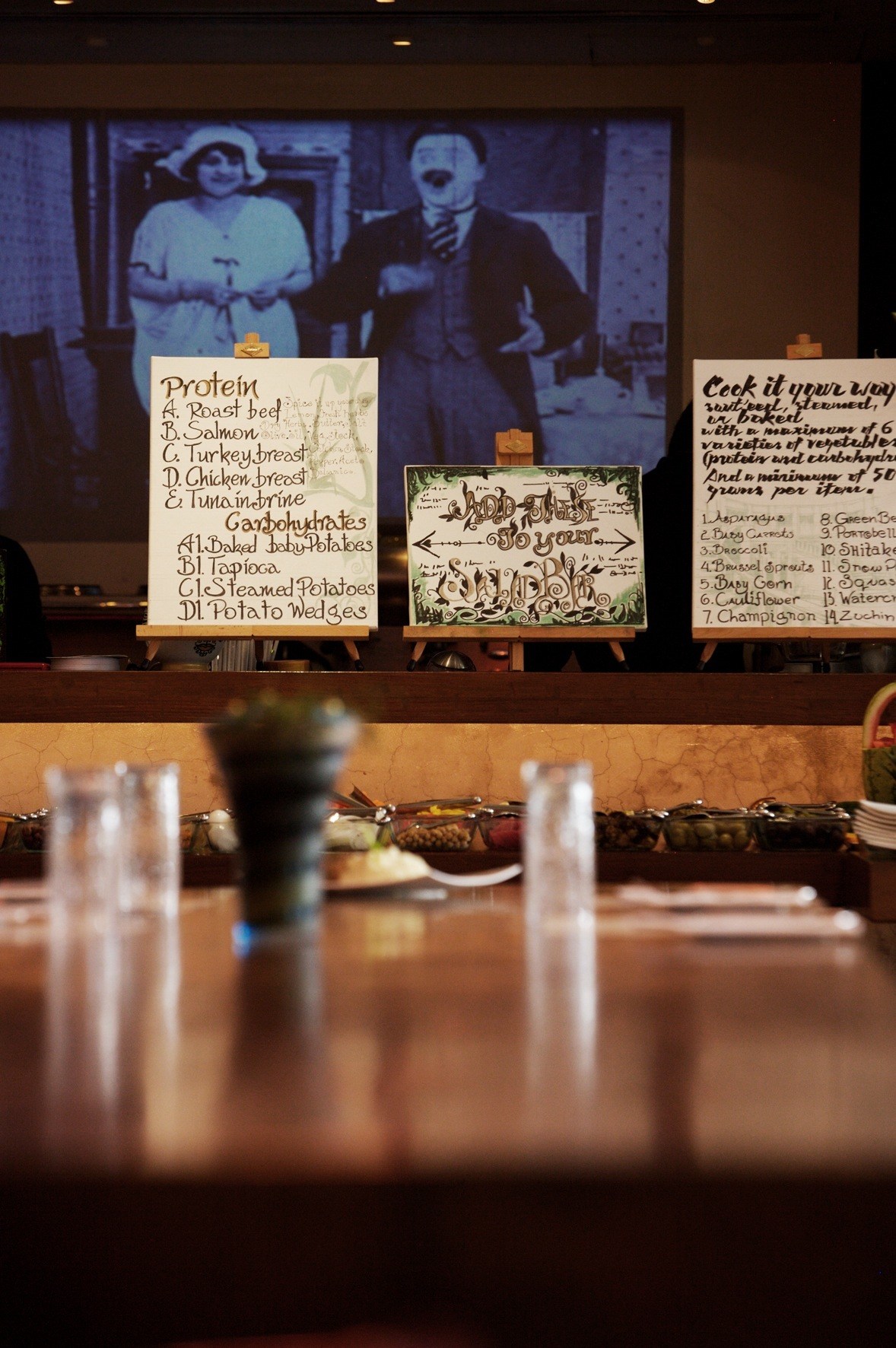 Saladicious restaurant's central table with handwritten menu boards and a Charlie Chaplin film still projected on the wall.