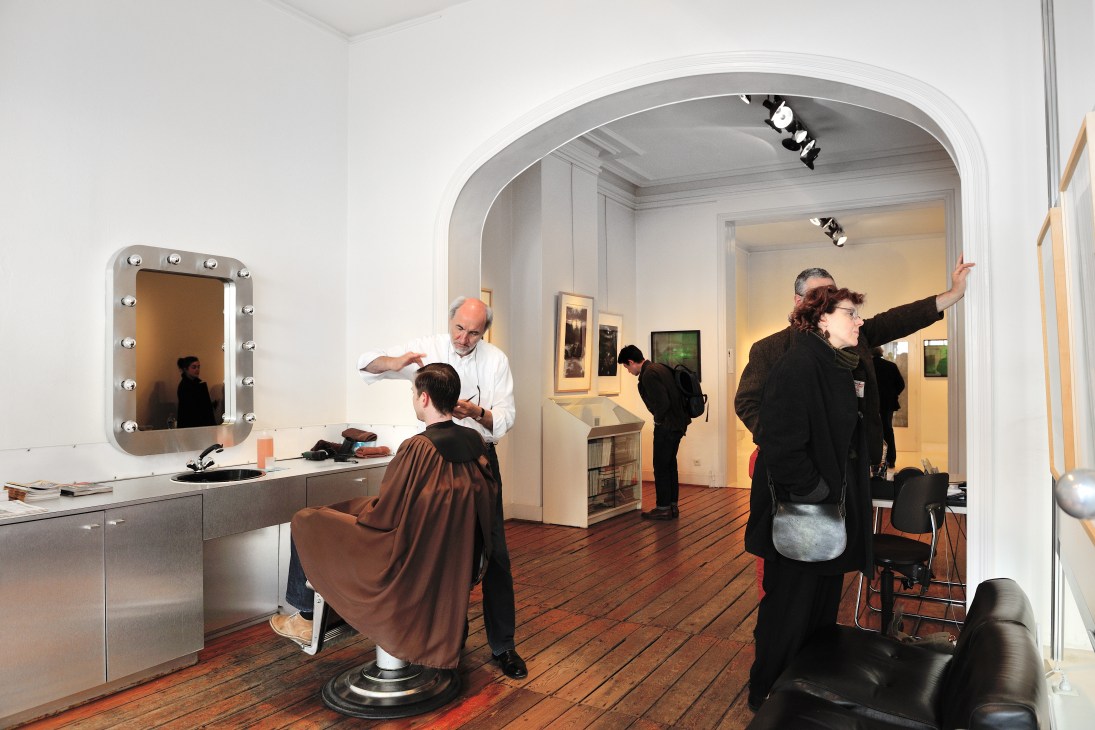 Interior of Le Salon d'Art et de Coiffure showing a hairdresser with client and gallery visitors viewing artworks.
