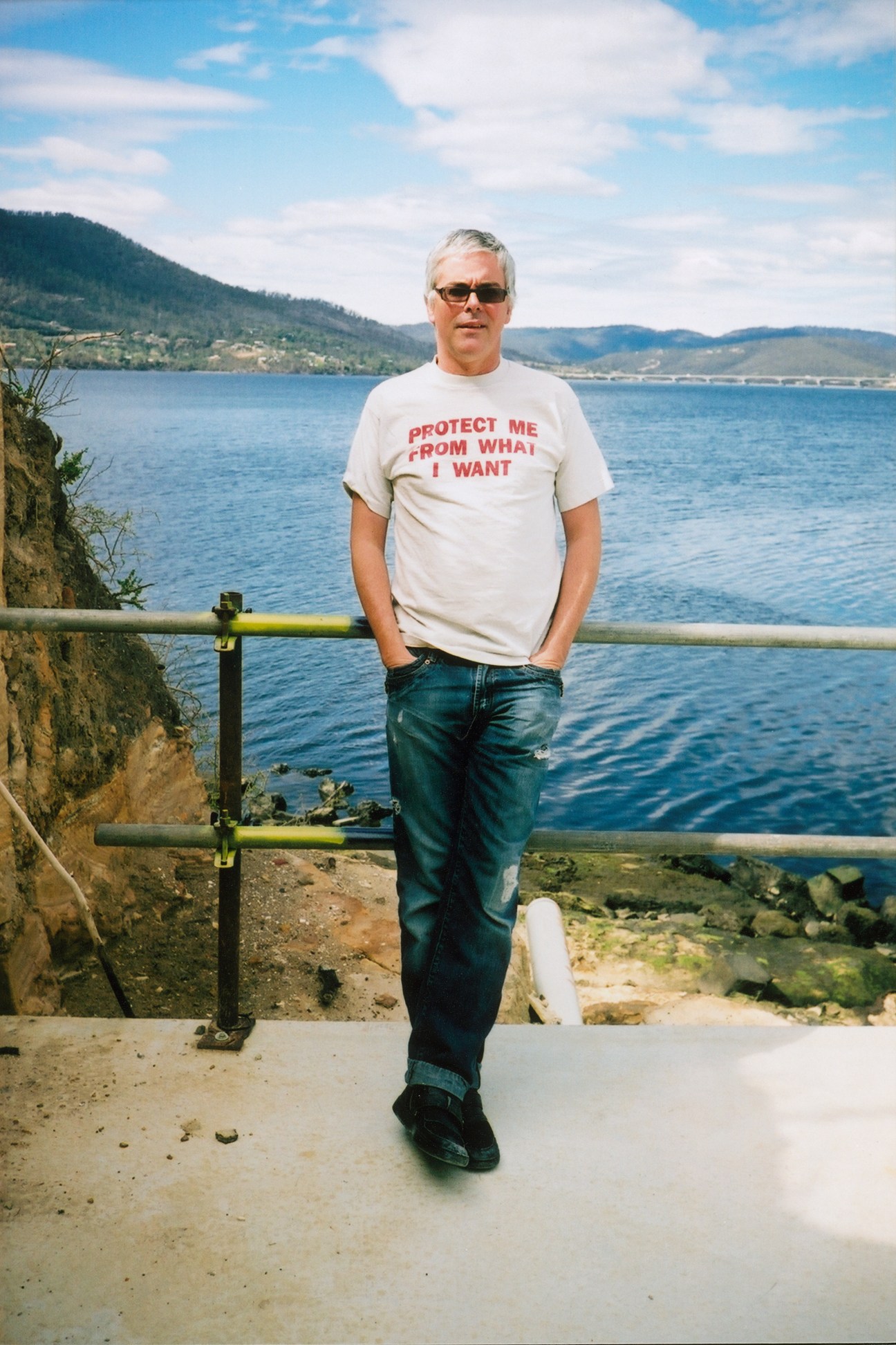 David Walsh, art collector and owner of MONA gallery, standing by a waterside railing wearing a graphic t-shirt.
