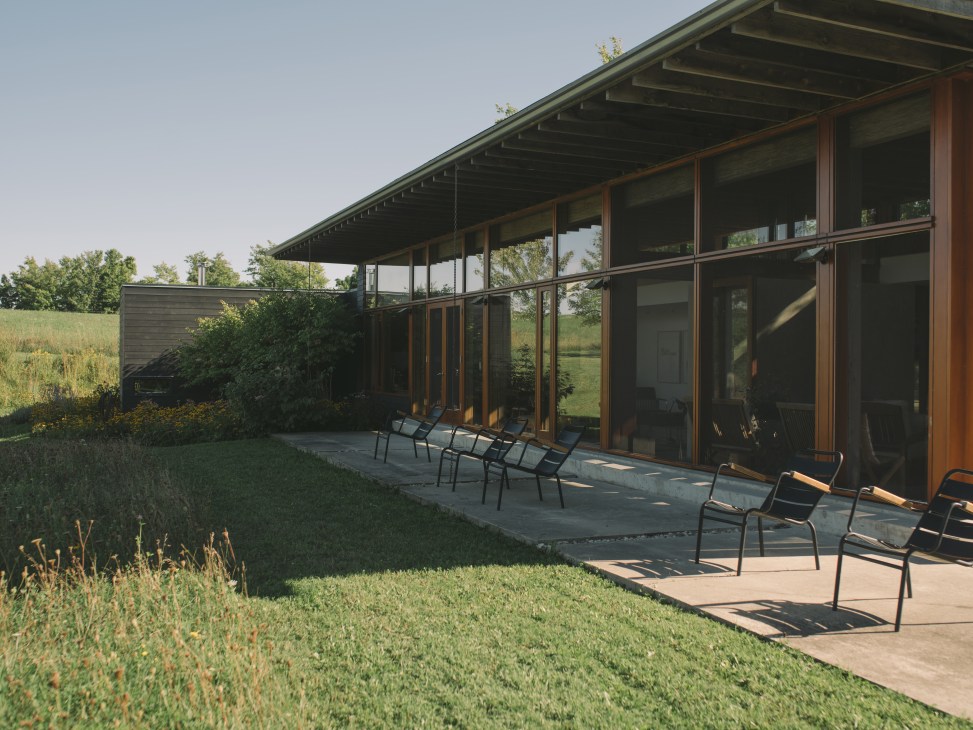 Modern house with glass walls, concrete terrace, and black outdoor chairs in rural landscape setting