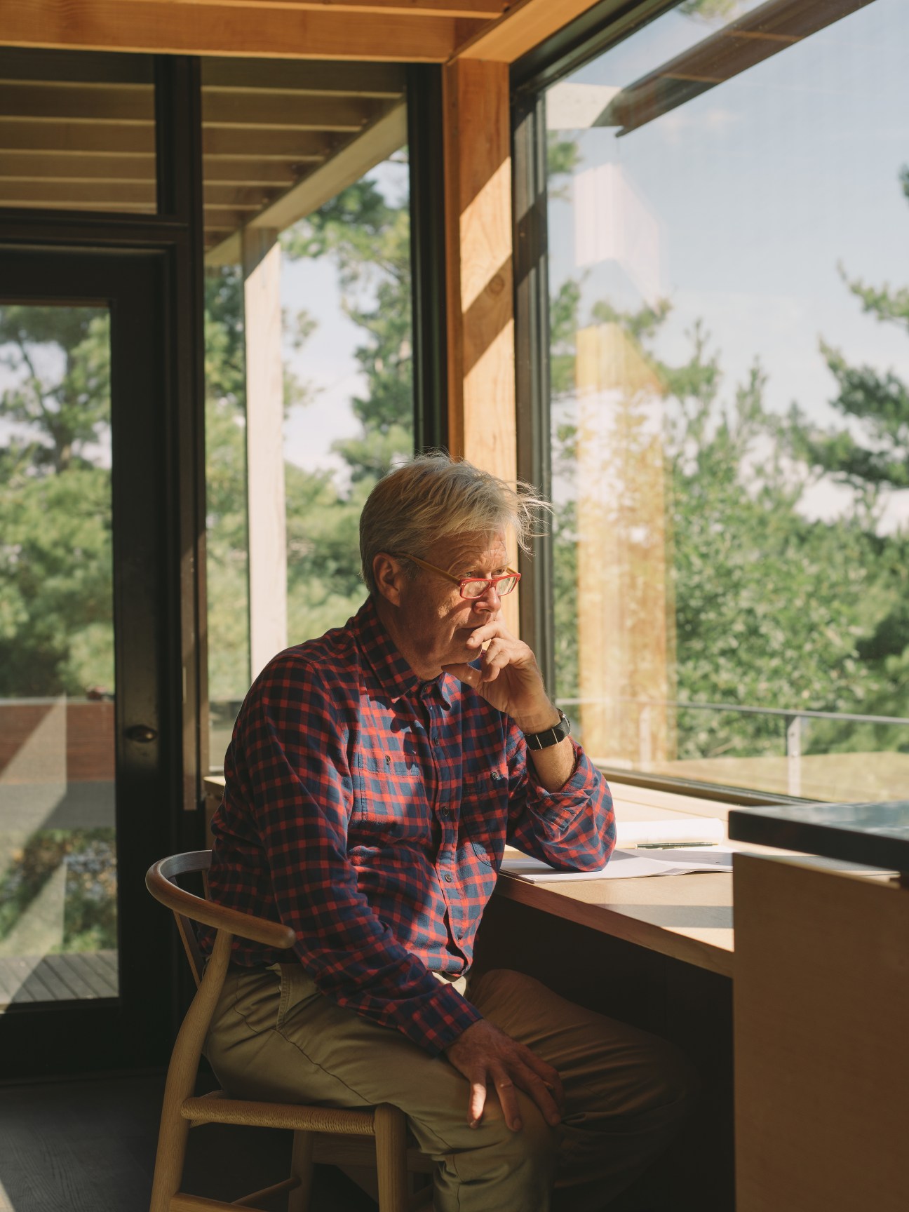 Elderly man in plaid shirt sitting at wooden desk in modern cabin with large windows overlooking forested landscape