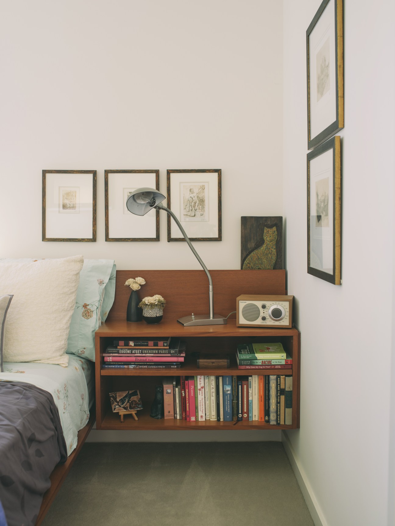 Bedroom with wooden floating nightstand containing books, desk lamp, radio, and framed artwork on white walls