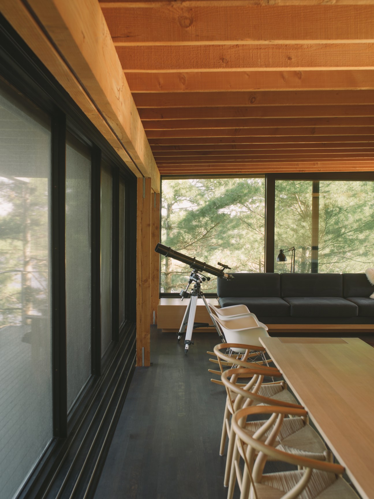 Modern living room with exposed wooden ceiling beams, telescope, dining table, and floor-to-ceiling windows