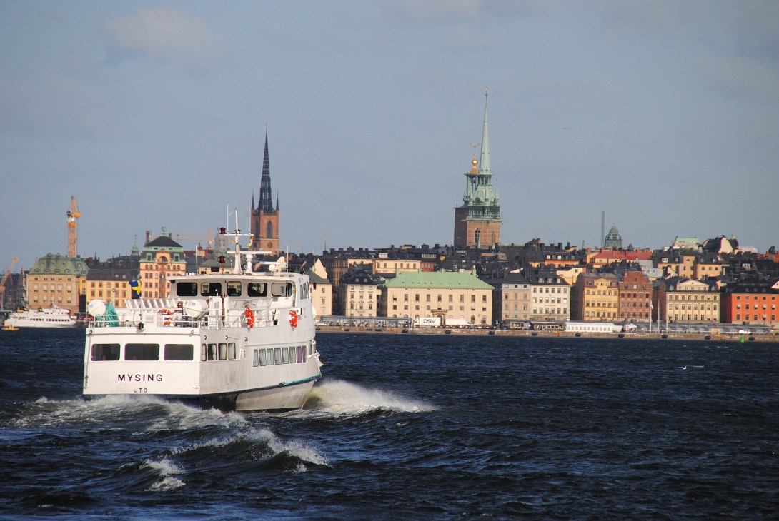White commuter ferry boat with Stockholm's colorful waterfront buildings and church spires in the background.
