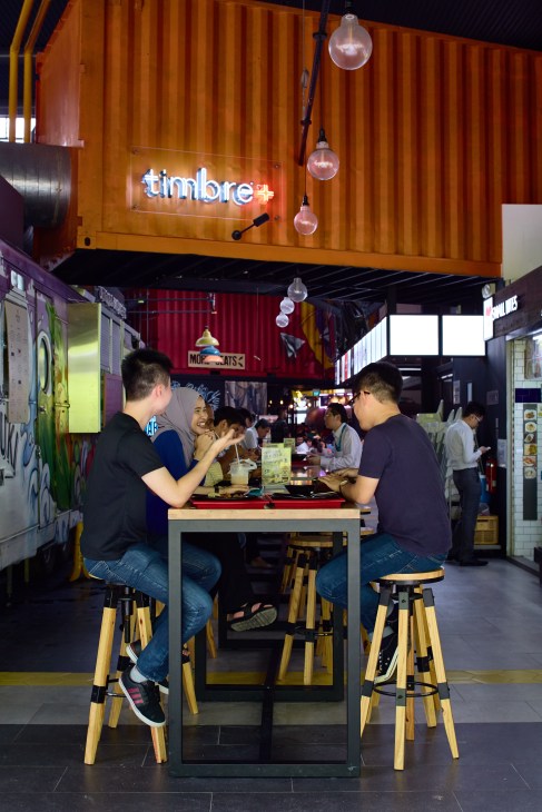 Diners enjoy food at a table in the Timbre+ food hall in Singapore, with the venue's neon sign visible above.