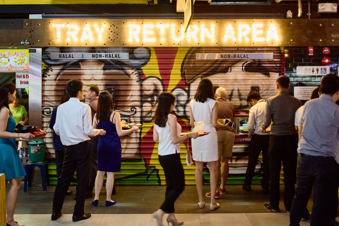 Diners with trays queue at a tray return area in a hawker centre food stall.