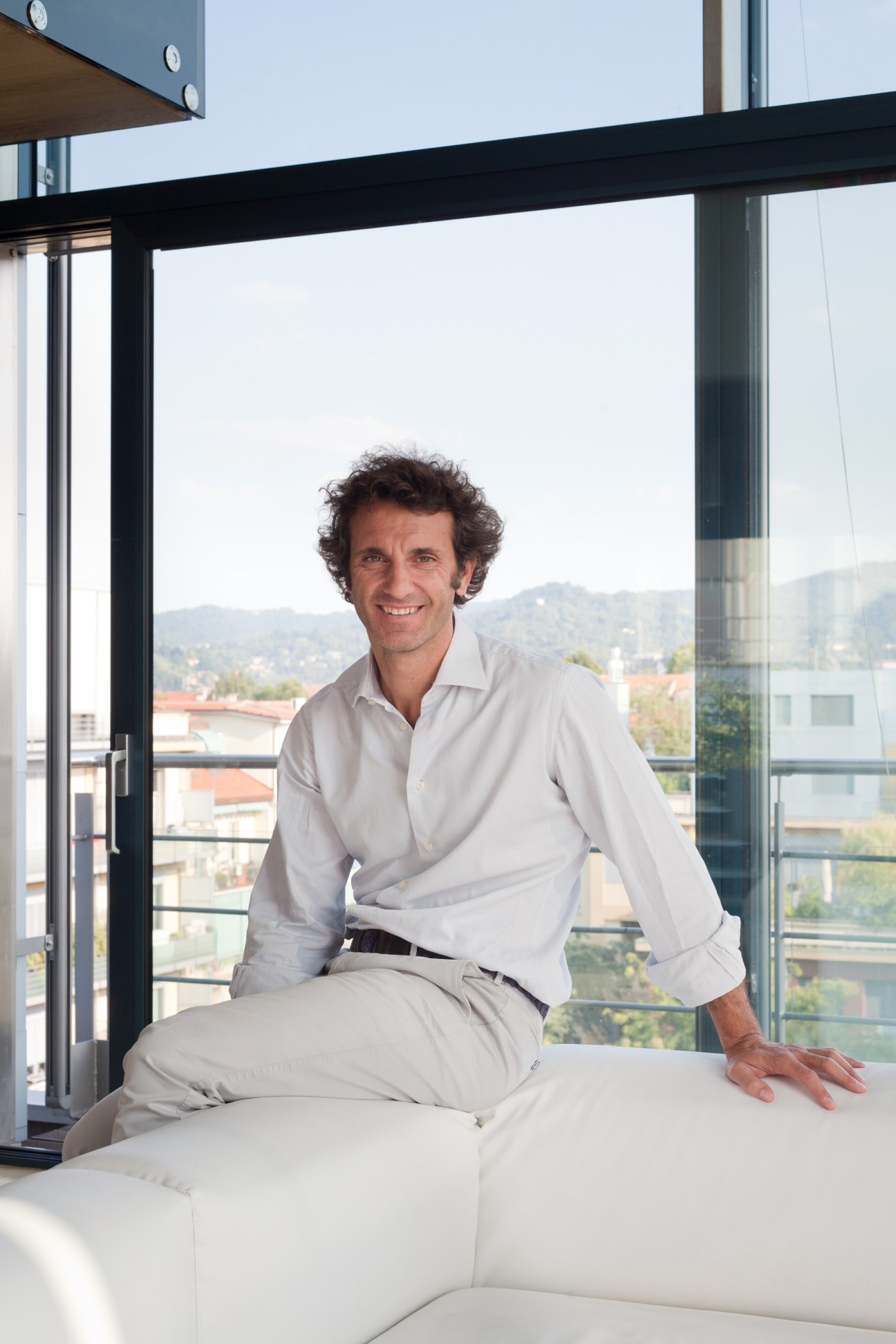 Architect Alberto Gallesio sits on a white sofa in a modern loft with floor-to-ceiling windows overlooking Turin.
