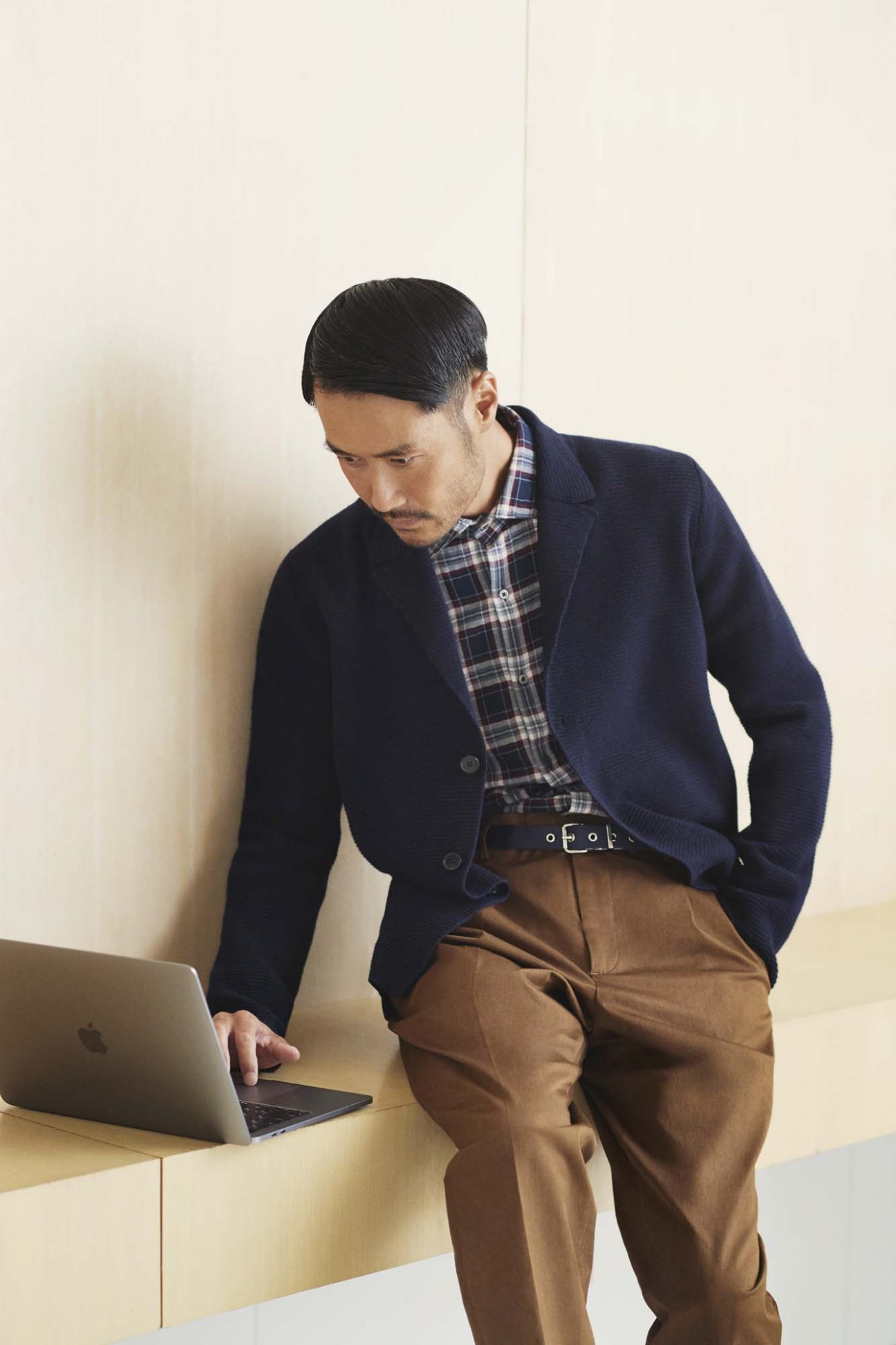 Man in layered business casual outfit with plaid shirt, navy blazer and brown trousers working at laptop.