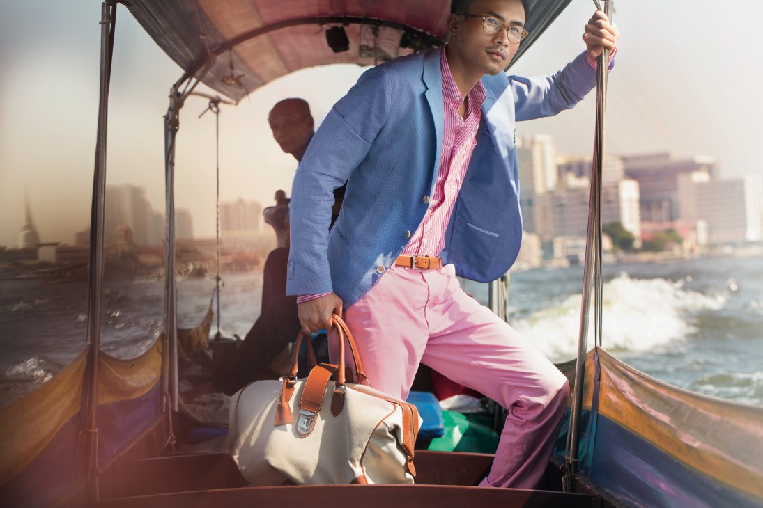 Man in blue jacket and pink trousers holding Hackett bag on a Bangkok river boat with city skyline behind.