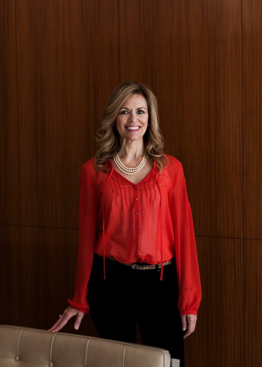 Woman in red blouse and black pants smiling at camera against wood paneled wall.