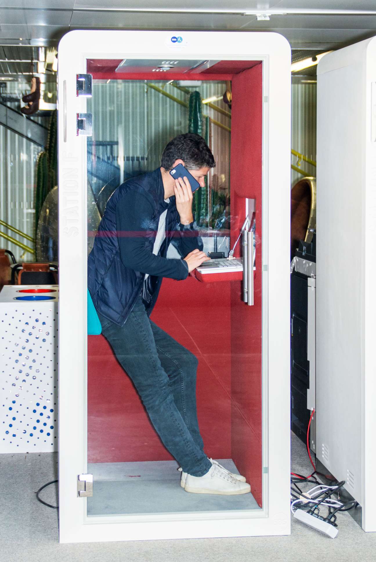 Man in soundproof booth at Station F co-working space working on laptop while on phone call.
