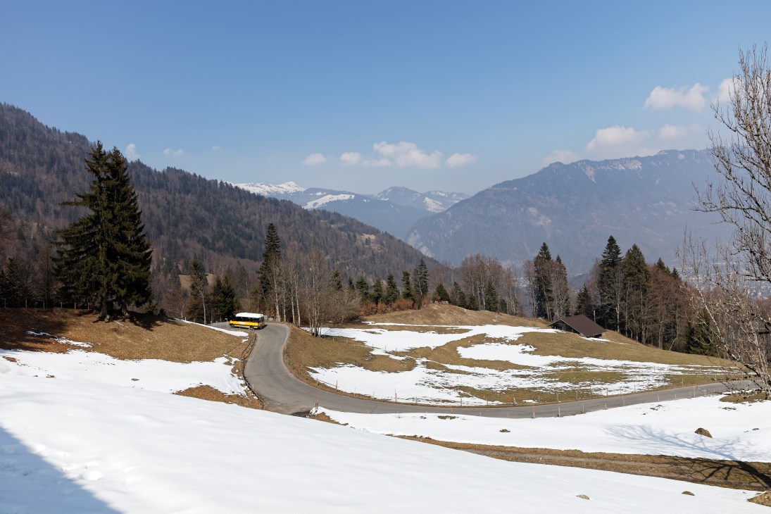 Yellow PostAuto bus navigating a winding mountain road through snowy Alpine terrain in the Bernese Oberland.