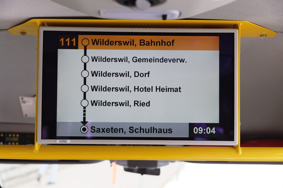 Digital display inside a yellow PostAuto bus showing route 111 stops including Saxeten, Schulhaus at 09:04.